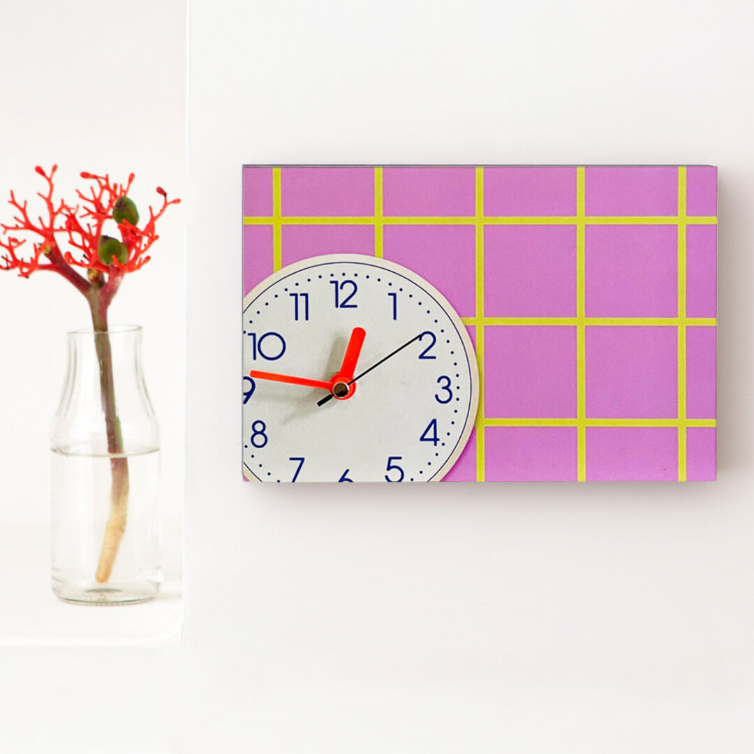 Wanduhr mit rosa Karomuster, gelben Linien und neonroten Zeigern auf weißem Zifferblatt