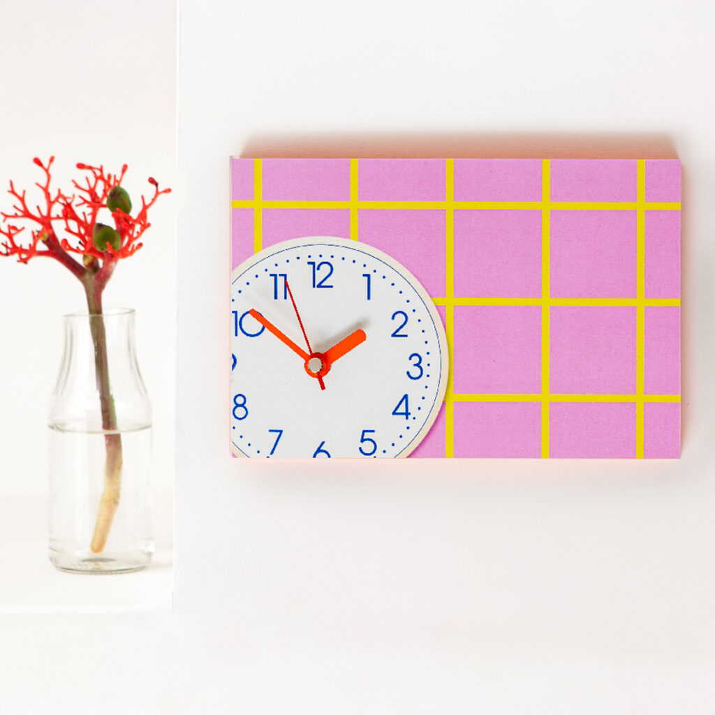 Wanduhr mit rosa Karomuster, gelben Linien und neonroten Zeigern auf weißem Zifferblatt und Neonrand