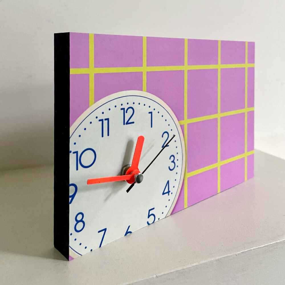 Wanduhr mit rosa Karomuster und gelben Linien und neonroten Zeigern auf weißem Zifferblatt