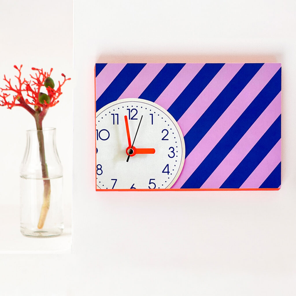 Wanduhr mit rosa, blauen Streifen und neonroten Zeigern auf weißem Zifferblatt und Neonrand